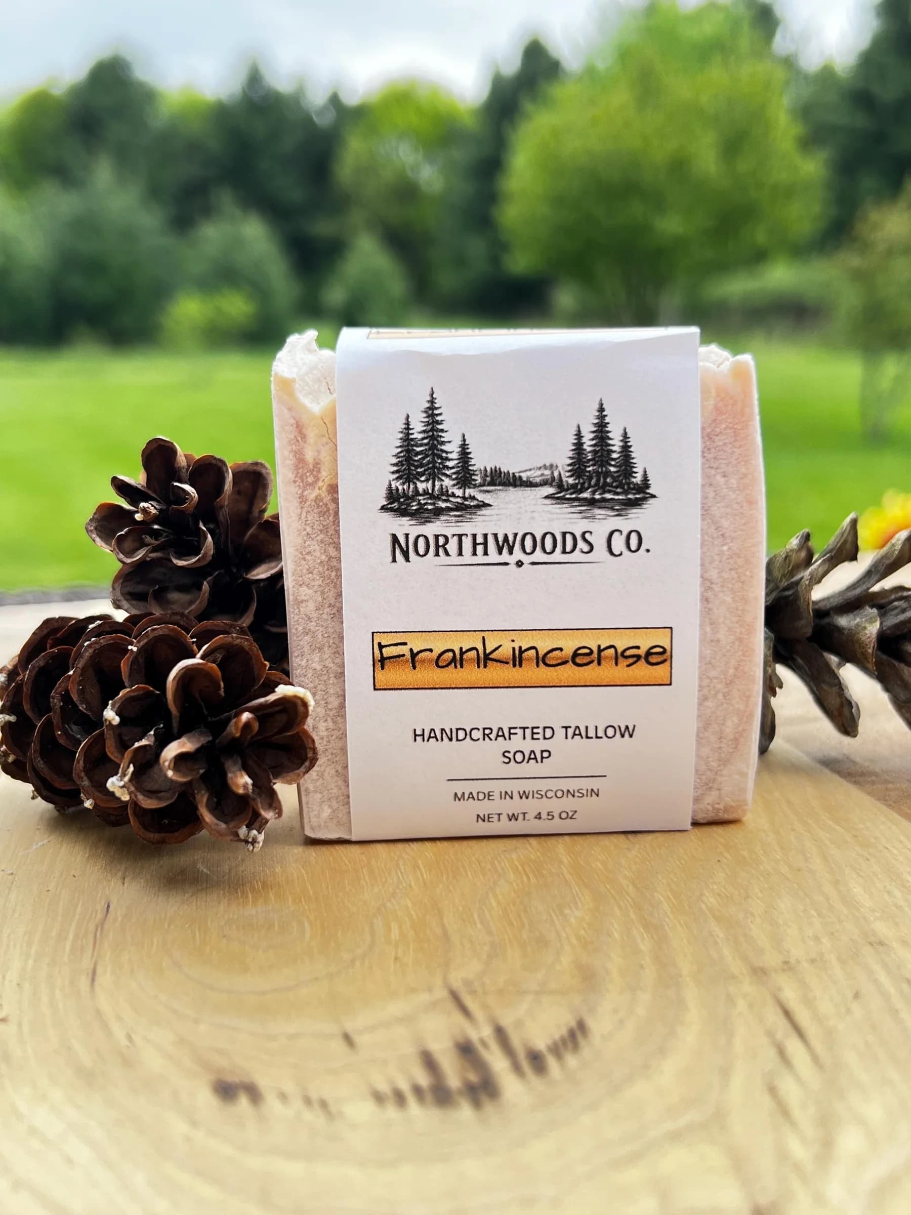 May include: A bar of Frankincense handcrafted tallow soap from Northwoods Co. is displayed with pine cones. The soap is wrapped in a white label with a forest scene and the text "Frankincense". The n