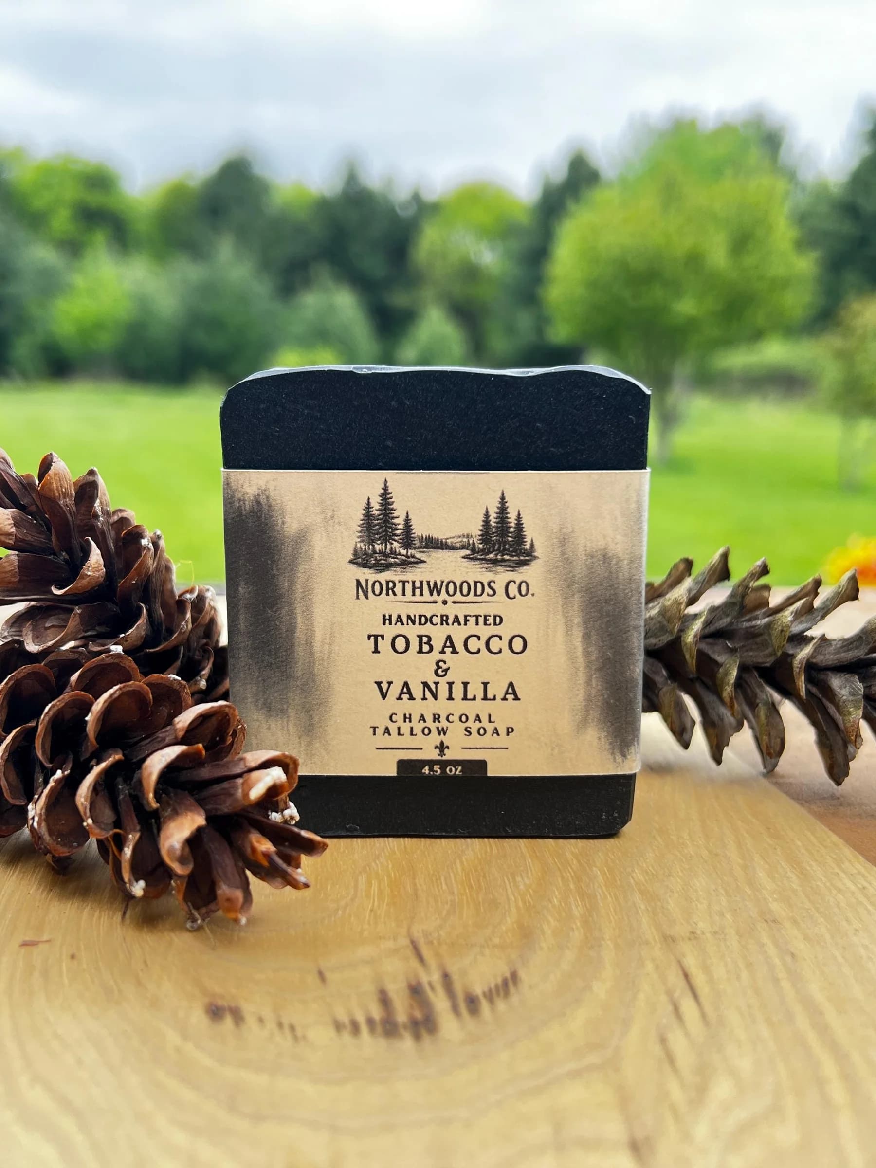 May include: A black, rectangular bar of charcoal tallow soap with a paper label. The label reads "Northwoods Co. Handcrafted Tobacco & Vanilla Charcoal Tallow Soap" and "4.5 oz". Two pine cones are o