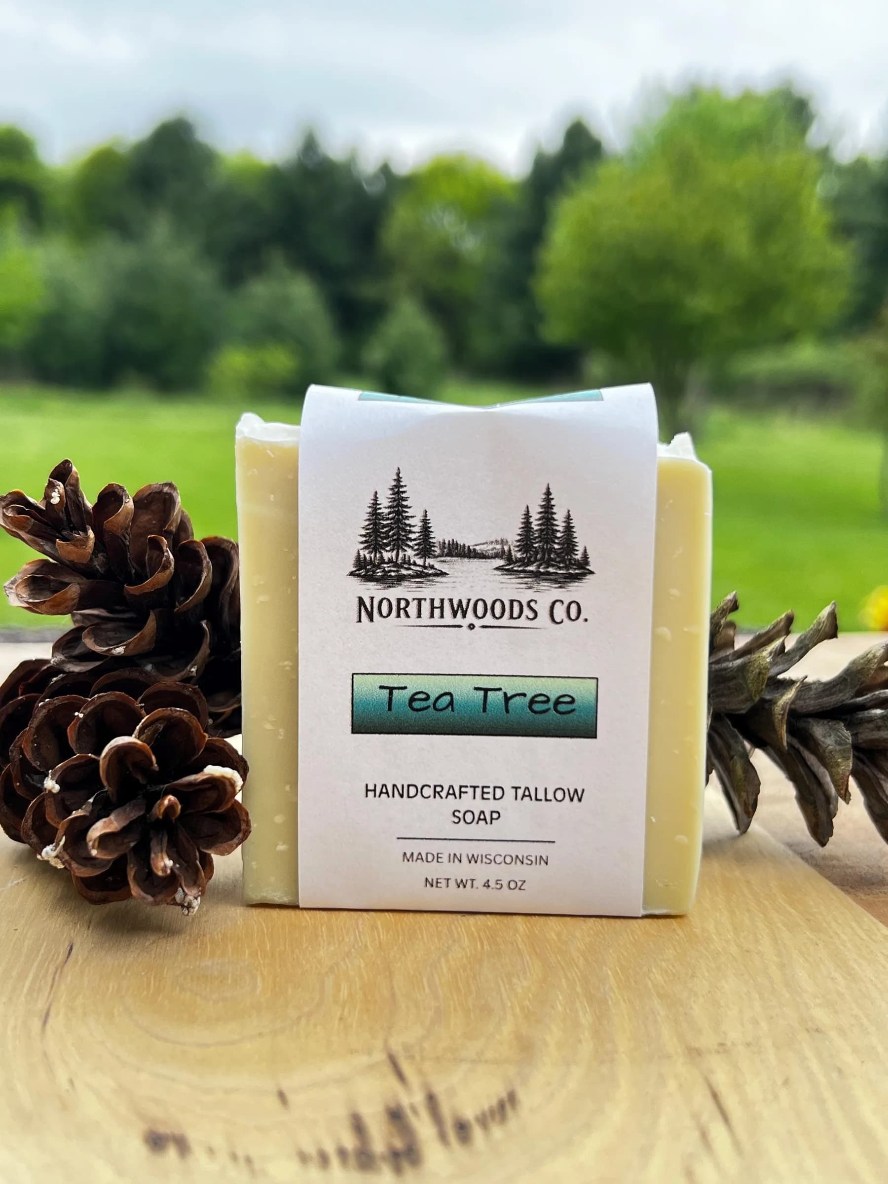 May include: A bar of handcrafted tallow soap with a label that reads "Tea Tree" and "Northwoods Co." The soap is light yellow and is next to two pine cones. The net weight is 4.5 oz.
