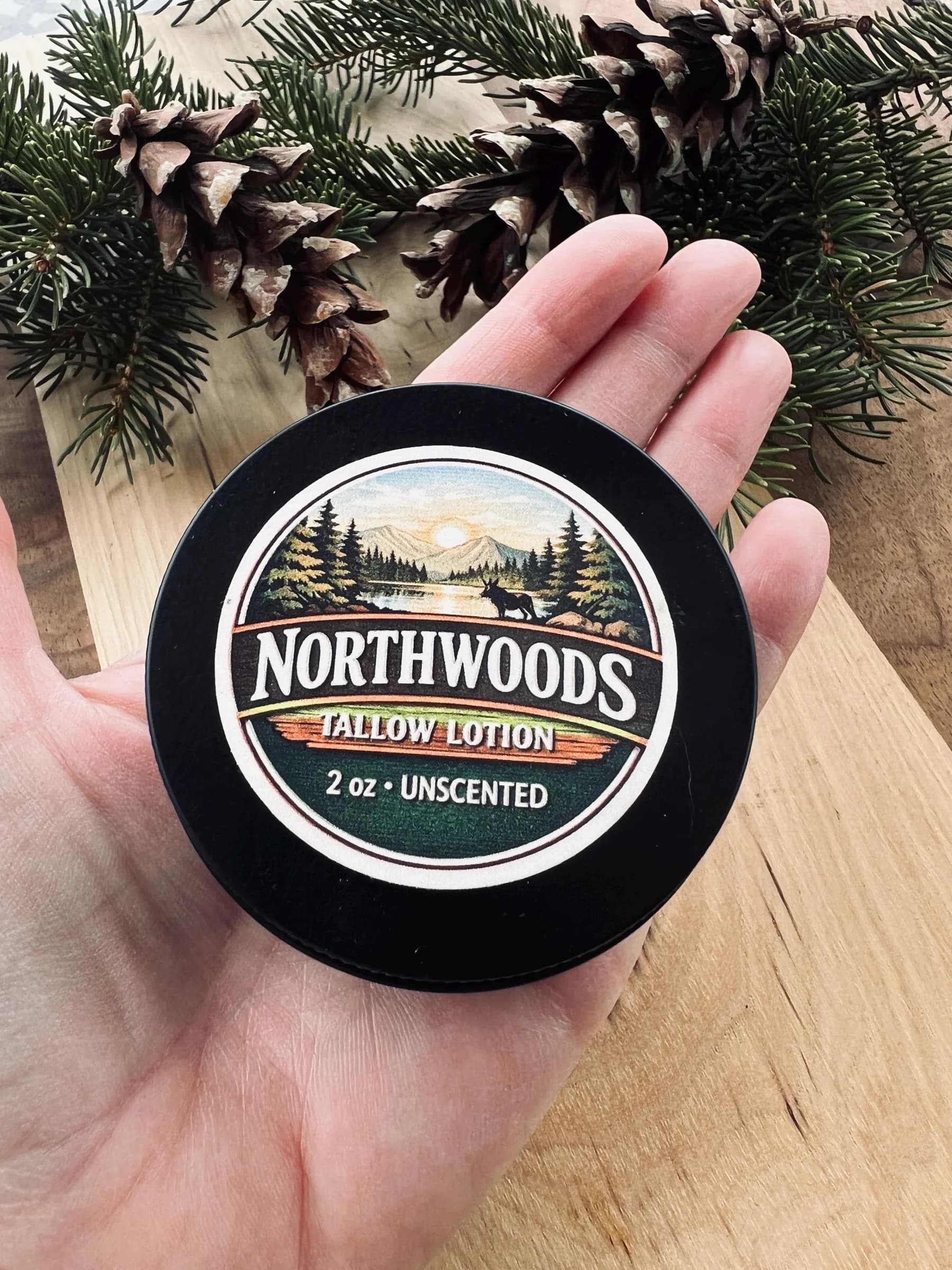 May include: A black, round container of Northwoods Tallow Lotion, 2 oz, unscented. The label features a landscape scene with trees, a lake, and a deer. The container is held in a person's hand, with