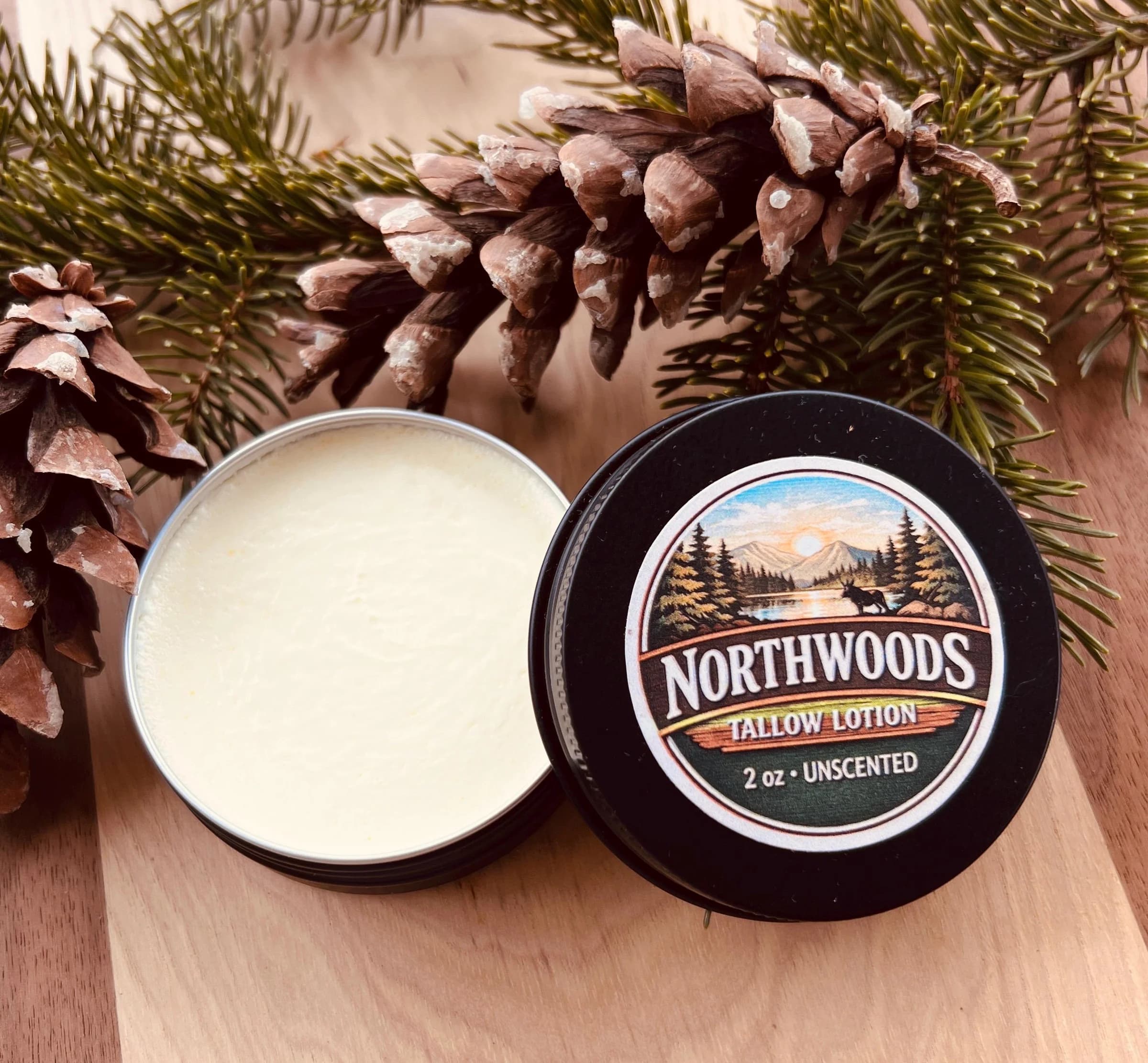 May include: An open, round, black tin of unscented tallow lotion next to a closed tin with a label that reads "Northwoods Tallow Lotion". The lotion is light yellow. Pine needles and pine cones are i